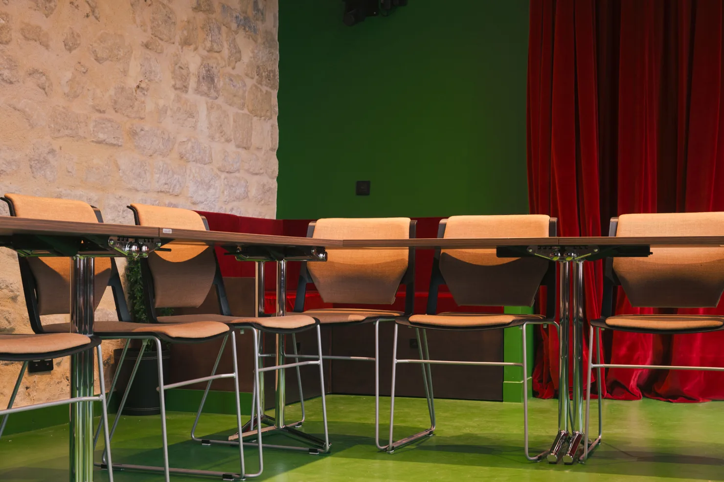 Une longue table et ses chaises claires, devant le rideau de velours rouge et le mur de pierre.