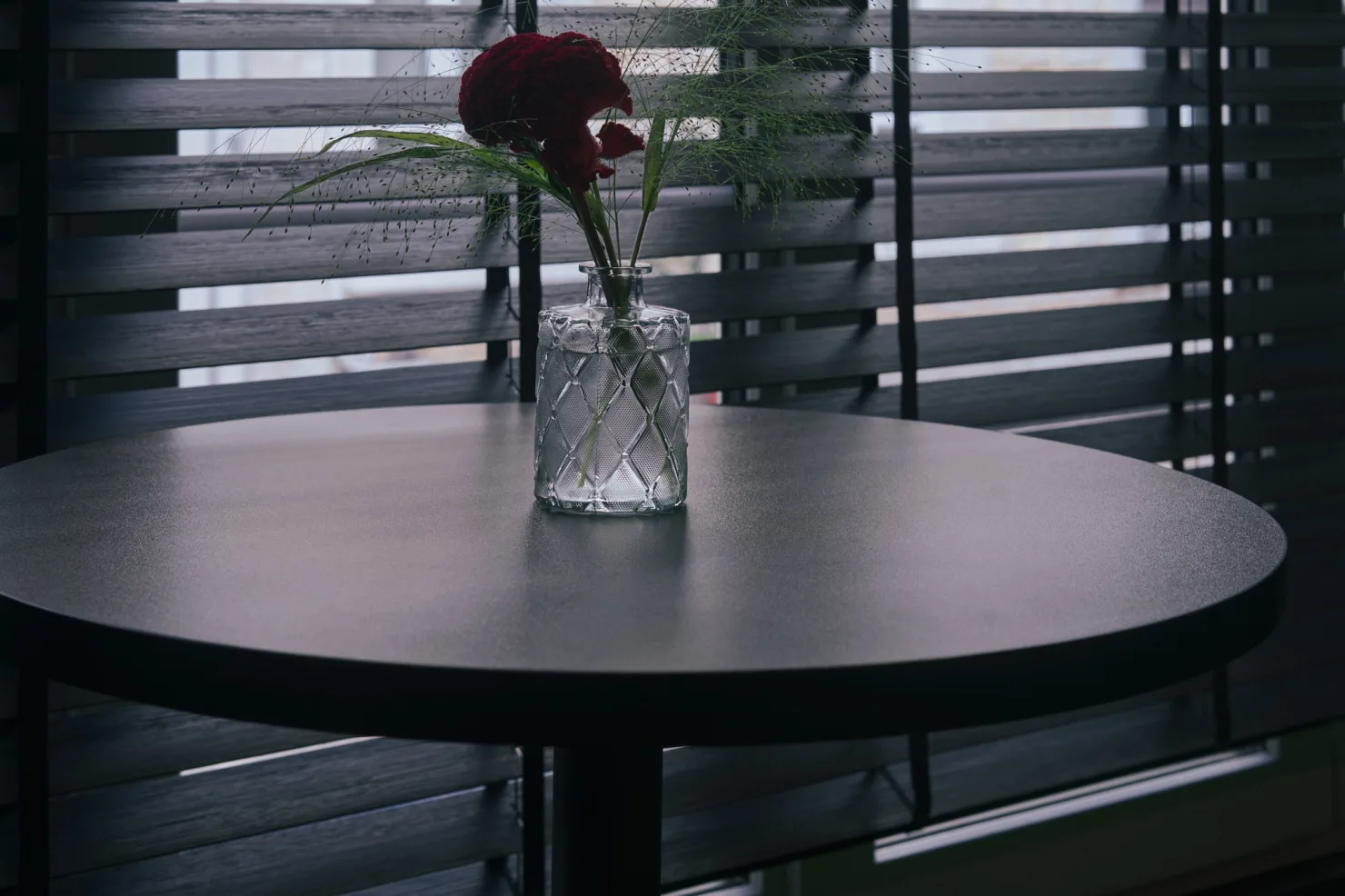 Le plateau d'un mange-debout noir et un bouquet de célosie dans un vase, devant les stores.