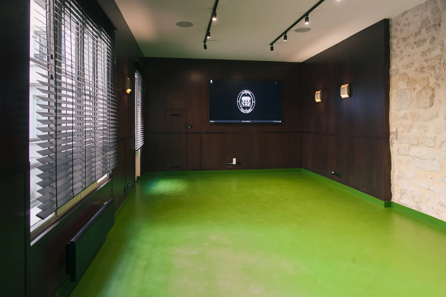 Le salon de réception vide : boiseries sombres, mur de pierre, sol vert et grandes fenêtres.